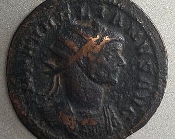May include: A dark brown, antique coin with a profile of a person wearing a crown. The coin has visible wear and tear, with some areas showing a copper color. The coin's edges are rounded, and the surface has a textured appearance.