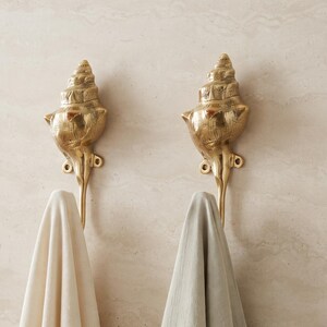 May include: Two gold-toned metal wall hooks shaped like conch shells, each featuring a mounting plate with two screw holes. A solid cream-colored towel hangs from the left hook, and a solid light gray towel hangs from the right hook.