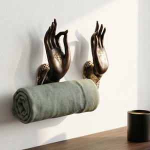 May include: Bronze-colored hand-shaped wall hooks holding a rolled-up green towel. The hooks are mounted on a white wall. A black mug sits on a wooden surface, suggesting a spa or bathroom setting.