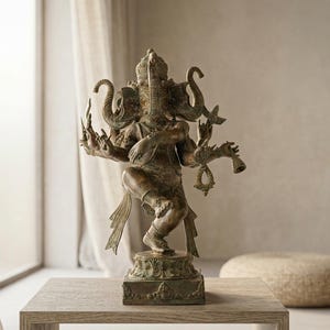 Op de afbeelding: Bronzen beeld van de hindoe&iuml;stische godheid Ganesha, met een olifantenkop en meerdere armen. Het beeld staat op een houten tafel. Een vaas met groen en een ronde poef zijn ook in de sc&egrave;ne, bij een raam.