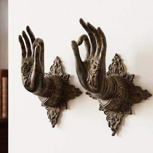 Puede incluir: Dos perchas de pared de color bronce con forma de manos en un gesto de bendición. Cada mano está montada sobre una base decorativa en forma de estrella. Las perchas se exhiben contra una pared blanca, lo que sugiere un tema decorativo o espiritual.