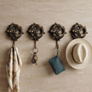 May include: Four circular bronze-toned wall hooks, each featuring a sculpted mermaid design with a curved tail. Each hook has a downward-facing metal prong. Items hanging from the hooks include a patterned scarf, a key ring, a small pouch, and a hat.
