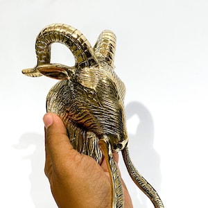 Puede incluir: Un llamador de puerta de latón dorado con forma de cabeza de carnero con cuernos detallados y una superficie texturizada.
