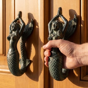 Op de afbeelding: Bronzen deurkloppers in de vorm van zeemeerminnen, bevestigd op een houten deur. De zeemeerminnen hebben hun handen boven hun hoofd gevouwen. De deur is gemaakt van lichtbruin hout. Een hand houdt een van de kloppers vast.