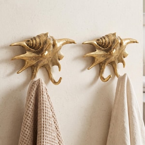 May include: Two gold-toned wall hooks shaped like spiky seashells. Each hook features a textured, metallic finish with a curved lower prong designed for hanging items. A beige waffle-weave towel hangs from the left hook, and a plain towel hangs from the right.