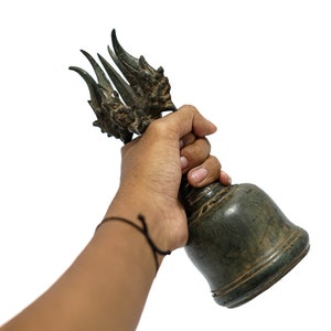 Puede incluir: Una mano sostiene una campana de bronce con una parte superior decorativa que presenta múltiples cabezas y garras de dragón. La campana es de color verde oscuro y tiene una superficie texturizada.