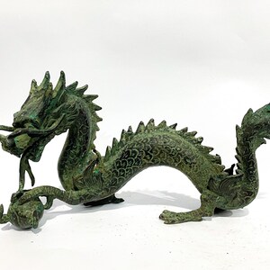 May include: A bronze-colored dragon sculpture with a verdigris finish. The dragon has a detailed, scaled body, a spiky dorsal ridge, and a curled tail. It holds a sphere in its claws and has an open mouth.