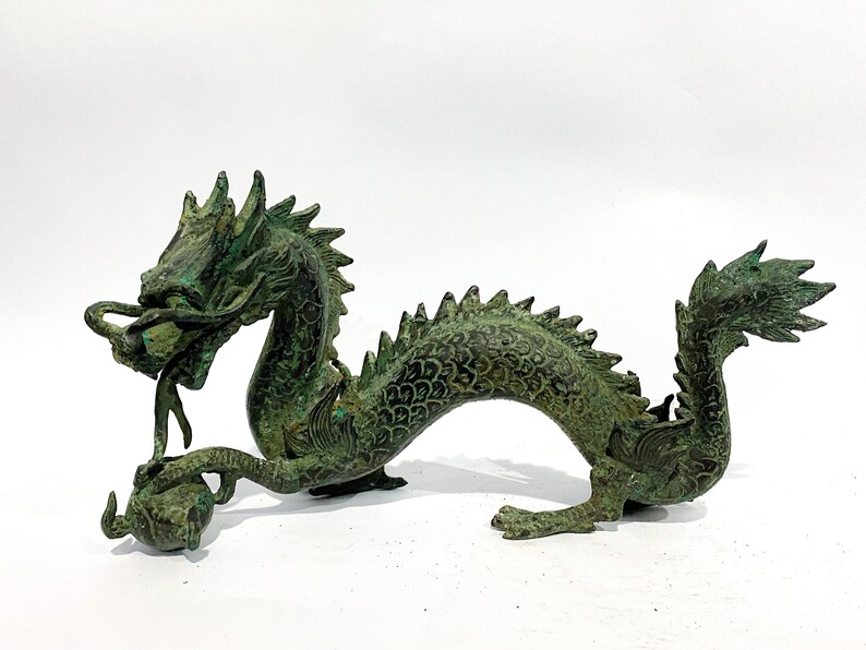 May include: A bronze-colored dragon sculpture with a verdigris finish. The dragon has a detailed, scaled body, a spiky dorsal ridge, and a curled tail. It holds a sphere in its claws and has an open mouth.