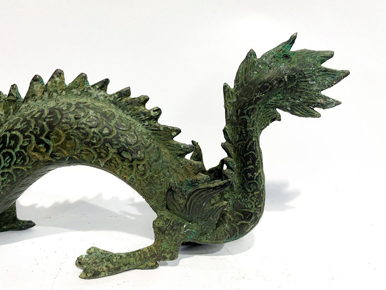 May include: A detailed bronze dragon sculpture with a verdigris finish. The dragon features intricate scale and fin details, with a curved body and a raised claw. The sculpture is a decorative piece, likely for home decor or display.