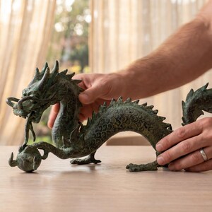 May include: A bronze-colored dragon sculpture with intricate details, holding a sphere in its mouth. The dragon has a textured surface and is positioned on a light-colored wooden table. The sculpture is being held by two hands.