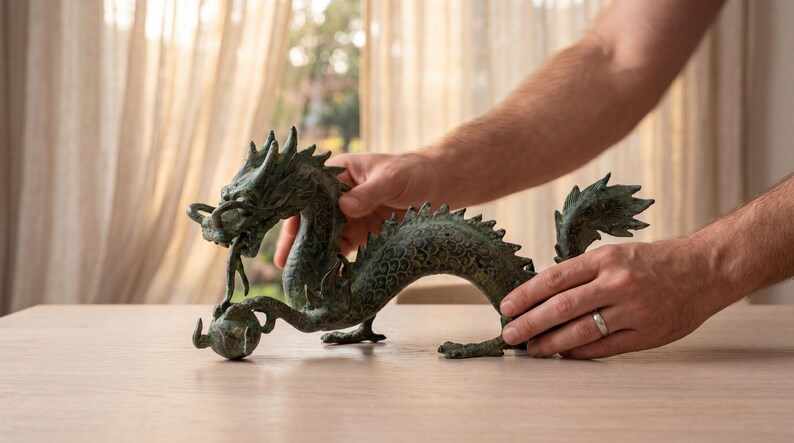 May include: A bronze-colored dragon sculpture with intricate details, holding a sphere in its mouth. The dragon has a textured surface and is positioned on a light-colored wooden table. The sculpture is being held by two hands.