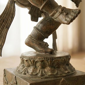 Op de afbeelding: Een bronzen metalen standbeeld van een dansende figuur met &eacute;&eacute;n been omhoog, versierd met gedetailleerde enkelbanden en vloeiende stoffen elementen. De figuur staat op een ronde lotusvormige voetstuk op een vierkante basis met patronen.