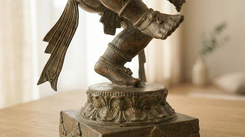 Op de afbeelding: Een bronzen metalen standbeeld van een dansende figuur met &eacute;&eacute;n been omhoog, versierd met gedetailleerde enkelbanden en vloeiende stoffen elementen. De figuur staat op een ronde lotusvormige voetstuk op een vierkante basis met patronen.