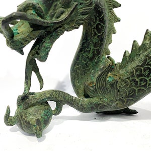 May include: A detailed bronze dragon sculpture with a weathered, verdigris finish. The dragon is depicted with an open mouth, intricate scales, and a curled tail, holding a sphere. The sculpture is set against a white background.