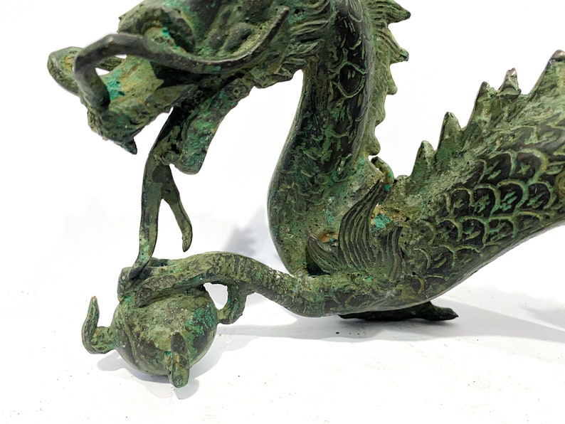 May include: A detailed bronze dragon sculpture with a weathered, verdigris finish. The dragon is depicted with an open mouth, intricate scales, and a curled tail, holding a sphere. The sculpture is set against a white background.