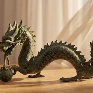 May include: A detailed bronze dragon figurine with a textured surface. The dragon has a long, serpentine body, spiky dorsal fins, and a detailed head with open mouth. The figurine is on a wooden surface.