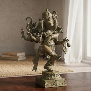 Op de afbeelding: Bronzen beeld van de hindoe&iuml;stische god Ganesha, met een olifantenkop en meerdere armen. Het beeld staat op een vierkante basis met gedetailleerde gravures. De achtergrond toont een raam met witte gordijnen, een tapijt en boeken.