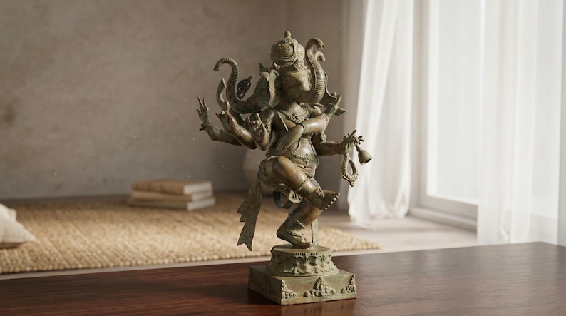Op de afbeelding: Bronzen beeld van de hindoe&iuml;stische god Ganesha, met een olifantenkop en meerdere armen. Het beeld staat op een vierkante basis met gedetailleerde gravures. De achtergrond toont een raam met witte gordijnen, een tapijt en boeken.