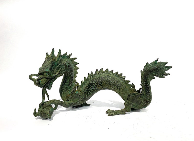 May include: A bronze-colored dragon figurine with a detailed, textured surface. The dragon has a long, serpentine body, a detailed head with a flowing mane, and a curled tail. The dragon is holding a sphere in its claws. The figurine is approximately 10 inches long.