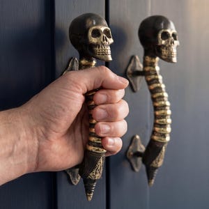 Puede incluir: Dos tiradores de puerta decorativos con diseños de calaveras. Los tiradores son negros con detalles dorados, sobre una puerta azul oscuro. Los tiradores con forma de calavera añaden un toque único y espeluznante.