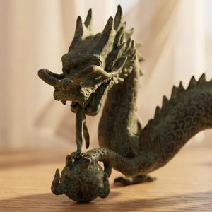 May include: A bronze dragon sculpture with intricate details. The dragon is depicted with a long body, spiky dorsal fins, and a detailed head. It is holding a sphere in its claws, a classic representation of power and wisdom.