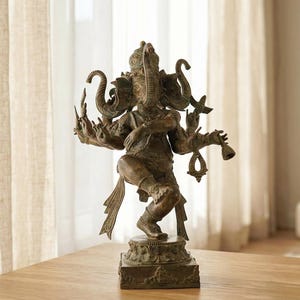 Op de afbeelding: Bronskleurig beeld van de hindoe&iuml;stische godheid Ganesha, met meerdere armen en een olifantenkop. Het beeld staat op een vierkante basis en op een houten tafel. De achtergrond omvat een transparant gordijn en een vaas.