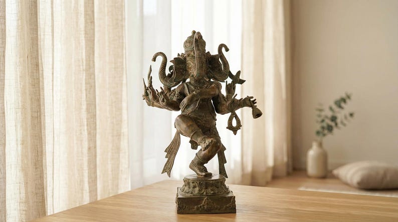 Op de afbeelding: Bronskleurig beeld van de hindoe&iuml;stische godheid Ganesha, met meerdere armen en een olifantenkop. Het beeld staat op een vierkante basis en op een houten tafel. De achtergrond omvat een transparant gordijn en een vaas.