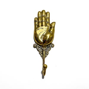 Pode incluir: Um gancho de metal dourado em forma de mão com detalhes ornamentados. O gancho é projetado para se assemelhar a uma mão humana com os dedos estendidos e um polegar apontando para o lado. O gancho é preso a uma base decorativa com um design de rolo.