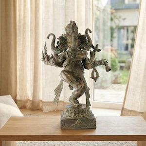 Op de afbeelding: Een bronskleurig standbeeld van de godheid Ganesha in een dansende houding met meerdere armen. De figuur heeft een olifantenkop, draagt versierde sieraden, houdt diverse symbolische voorwerpen vast en staat op een vierkante voet.