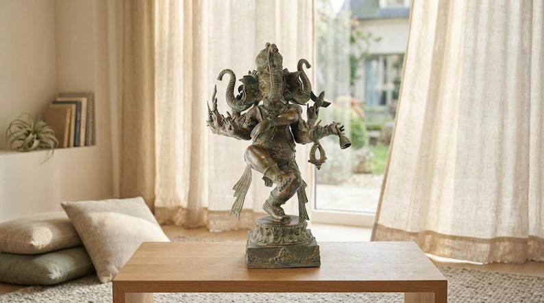 Op de afbeelding: Een bronskleurig standbeeld van de godheid Ganesha in een dansende houding met meerdere armen. De figuur heeft een olifantenkop, draagt versierde sieraden, houdt diverse symbolische voorwerpen vast en staat op een vierkante voet.