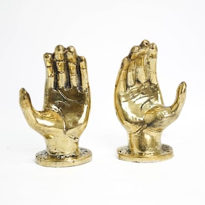 May include: Two gold-colored metal hand sculptures with open palms facing upwards. The sculptures are on a white surface.