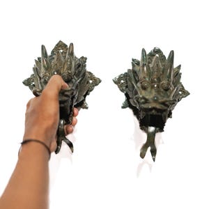 May include: Two bronze dragon head wall sconces. One is held by a hand. The sconces have detailed features, including sharp teeth, spiky manes, and clawed feet. The sconces are mounted on a decorative base with an ornate design.
