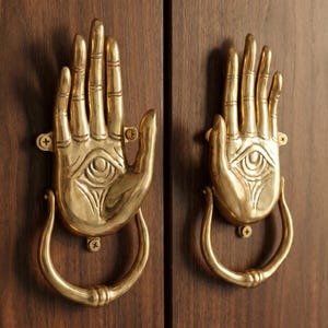 May include: Two brass hand-shaped door knockers with an eye design, mounted on a dark wood door. The knockers have a detailed, open-palm design with a curved handle below.