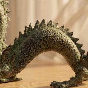 May include: A detailed bronze dragon sculpture with a textured surface. The dragon is in a curved pose, with its tail raised and its body arched. The sculpture is on a wooden surface.