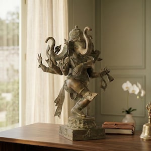 Op de afbeelding: Bronzen beeld van de hindoe-god Ganesha met meerdere armen, staand op een vierkante basis. Het beeld staat op een houten tafel, met een bel en een klein boekje in de buurt. De achtergrond omvat een raam en een vaas met witte bloemen.