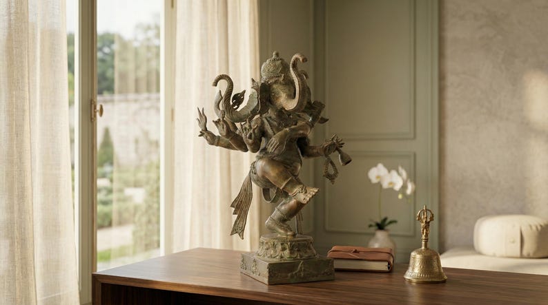 Op de afbeelding: Bronzen beeld van de hindoe-god Ganesha met meerdere armen, staand op een vierkante basis. Het beeld staat op een houten tafel, met een bel en een klein boekje in de buurt. De achtergrond omvat een raam en een vaas met witte bloemen.