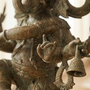 Op de afbeelding: Bronzen beeld van Ganesha, de hindoe&iuml;stische god. De sculptuur heeft meerdere armen, een olifantenkop en ingewikkelde details. E&eacute;n hand houdt een bel vast en een andere toont een gebaar. Het beeld heeft een verweerde, antieke afwerking.