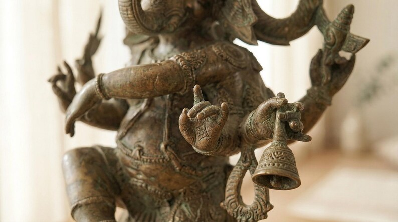 Op de afbeelding: Bronzen beeld van Ganesha, de hindoe&iuml;stische god. De sculptuur heeft meerdere armen, een olifantenkop en ingewikkelde details. E&eacute;n hand houdt een bel vast en een andere toont een gebaar. Het beeld heeft een verweerde, antieke afwerking.