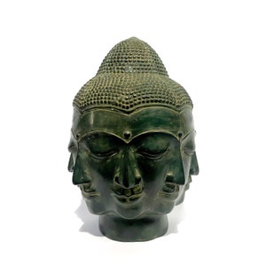 May include: A dark green Buddha head sculpture with multiple faces, each with closed eyes. The sculpture has a textured, detailed surface and a serene expression. The head is set against a white background.