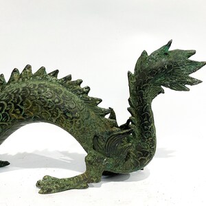 May include: A detailed bronze dragon sculpture with a verdigris finish. The dragon features intricate scales, a spiky dorsal ridge, and a curled tail. The sculpture is set against a white background, highlighting its detailed craftsmanship.
