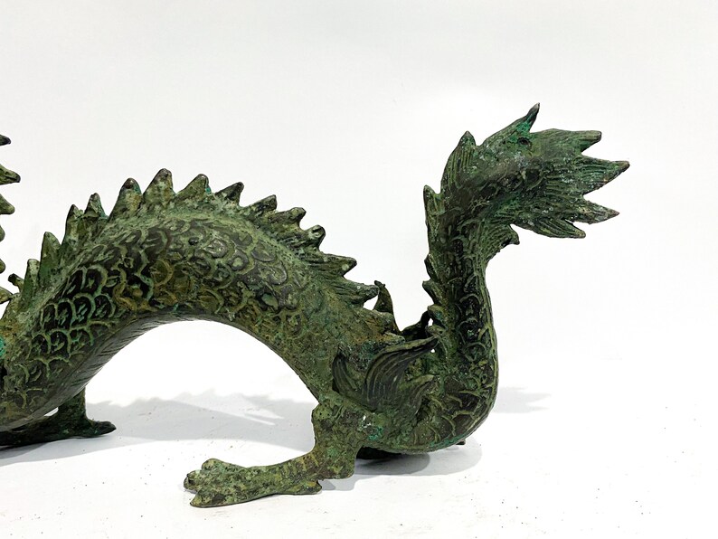 May include: A detailed bronze dragon sculpture with a verdigris finish. The dragon features intricate scales, a spiky dorsal ridge, and a curled tail. The sculpture is set against a white background, highlighting its detailed craftsmanship.