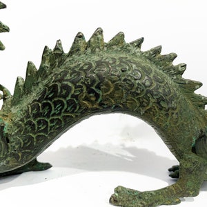 May include: A detailed bronze dragon sculpture with a weathered, verdigris finish. The dragon features intricate scale patterns, a spiky dorsal ridge, and clawed feet. The sculpture is set against a white background.