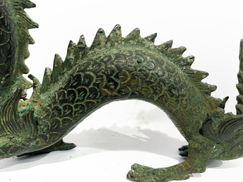 May include: A detailed bronze dragon sculpture with a weathered, verdigris finish. The dragon features intricate scale patterns, a spiky dorsal ridge, and clawed feet. The sculpture is set against a white background.