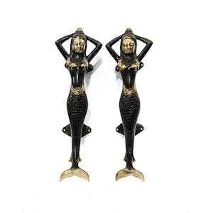 May include: Two bronze mermaid door handles. Each handle features a mermaid with her arms raised above her head. The mermaids have gold-coloured faces, chests, and tail fins. The handles are dark bronze with a decorative scale pattern.