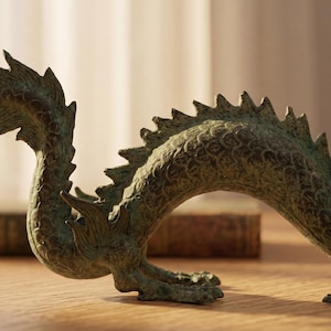 May include: A bronze-colored dragon figurine with intricate details, including scales and a spiky spine. The dragon is posed in a curved position, with its head and tail raised. The figurine is on a wooden surface.
