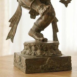 Op de afbeelding: Een bronskleurig metalen standbeeld van een dansende figuur op een cirkelvormig lotusvormig voetstuk op een vierkante basis. De figuur heeft gedetailleerde benen en voeten met uitlopende stoffen elementen vanaf de taille.