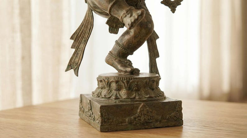 Op de afbeelding: Een bronskleurig metalen standbeeld van een dansende figuur op een cirkelvormig lotusvormig voetstuk op een vierkante basis. De figuur heeft gedetailleerde benen en voeten met uitlopende stoffen elementen vanaf de taille.