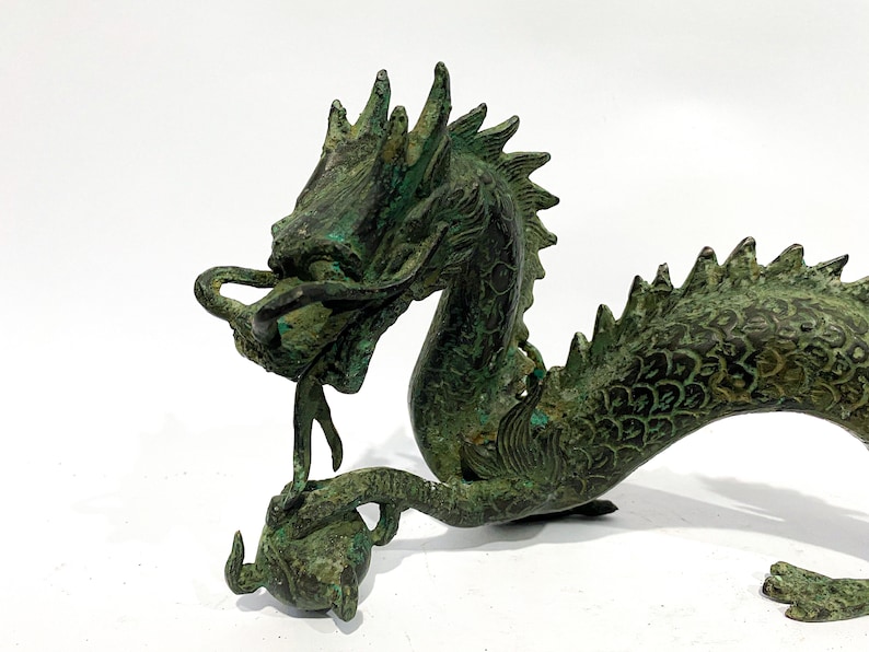 May include: A detailed bronze dragon sculpture with a verdigris finish. The dragon is depicted with intricate scales, a flowing mane, and a curled tail. It is holding a sphere in its claw, showcasing a traditional Asian art style.