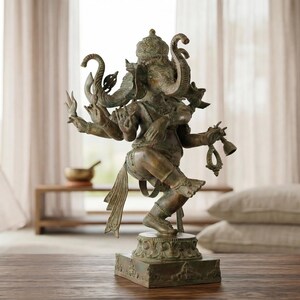 Op de afbeelding: Een bronskleurig standbeeld van de godheid Ganesha in een dansende houding met meerdere armen. De figuur heeft een olifantenkop, een kroon en ingewikkelde versieringen en staat op een vierkante voet met gedetailleerd houtsnijwerk.