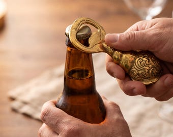 May include: A gold-colored bottle opener with an ornate design is used to open a brown beer bottle. The opener is held in place with one hand while the other hand holds the bottle. The scene is set on a wooden surface.